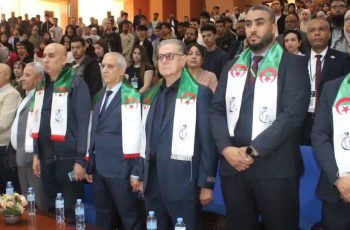 Group of men in suits standing at a formal ceremony, wearing white scarves with green and red Algerian flags; audience seated behind.