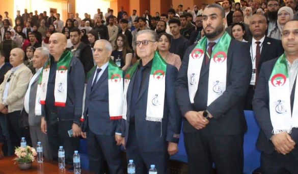 Group of men in suits standing at a formal ceremony, wearing white scarves with green and red Algerian flags; audience seated behind.