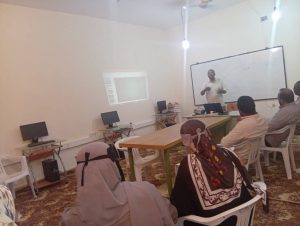 C0C86FD0-6223-441B-87C1-54421AC68CE4 - جامعة سبها | Sebha University Instructor presents near a whiteboard to a small class in a computer lab with idle monitors on desks behind them.