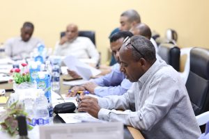 C1650947-1BC8-4F37-82FB-291D80343EFE - جامعة سبها | Sebha University Man in the foreground writing at a conference table, with other attendees blurred in the background of a meeting room.