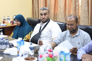 C38AA56A-98E8-461C-8234-82538AC32E4E - جامعة سبها | Sebha University Three adults seated around a conference table with drinks and snacks, facing the camera; man in white shirt in the center, woman in teal hijab on the left, man with glasses on the right.