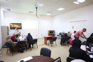 CF4324C3-2BDA-417A-949B-747D76FD0D52 - جامعة سبها | Sebha University Group of people in hijabs sitting around tables in a bright classroom with a ceiling projector and a screen at the front.