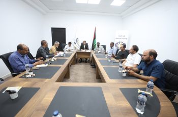 A group of men seated around a long conference table in a bright white room, with water bottles and snacks, engaged in a formal meeting.