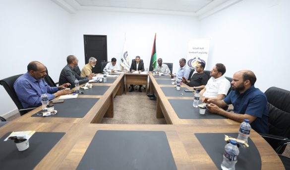 A group of men seated around a long conference table in a bright white room, with water bottles and snacks, engaged in a formal meeting.
