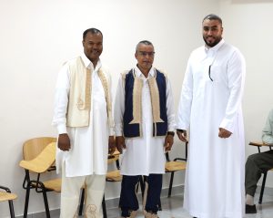 Three men wearing traditional white robes and embroidered vests pose for a photo in a classroom.','Older man in a blue-beige embroidered vest stands between two men in white thobes, in a classroom.','Group portrait of three men in traditional attire standing beside classroom chairs in a bright room.','Three men in cultural garments pose together with light walls and classroom chairs in the background.','Men in ceremonial robes stand together for a photo in a classroom setting.
