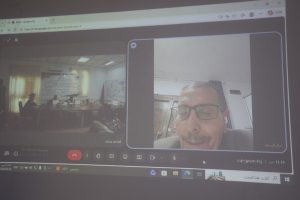 Google Meet video call on a computer screen, showing a man wearing glasses in the foreground and a conference room in the left window.