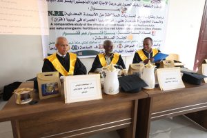 Three men in black gowns with gold sashes sit at a table in front of a large banner about a fertilizer study; teapots and gift boxes are on the table.