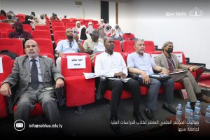 D947A330-4F7F-48D1-BC54-C11E134C674C - جامعة سبها | Sebha University Audience seated in red theater chairs at a university conference; front row includes men in suits and a masked attendee, Sabha University logo visible in the corner.