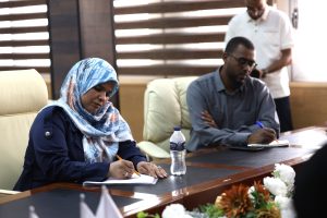 Woman in a blue and floral hijab writes on a notepad at a conference table, a water bottle in the foreground and a man in glasses writing nearby.
