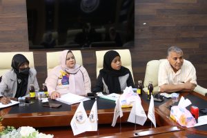 Four adults seated at a conference table in a wood-paneled room, with hijab-wearing women and a man on the right, beverages and papers on the table.