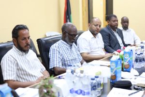 DC241EE7-7AA6-413E-8CDB-D6972EBA7B12 - جامعة سبها | Sebha University Men seated at a conference table during a meeting with juice boxes and water bottles on the table in front of them, appears to be a formal business discussion.