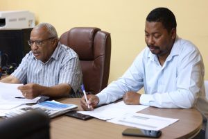 DC5FDC7F-1FF1-4425-963B-1880A1F00943 - جامعة سبها | Sebha University Two men sit at a desk reviewing documents in an office setting behind them are chairs and papers on the table.