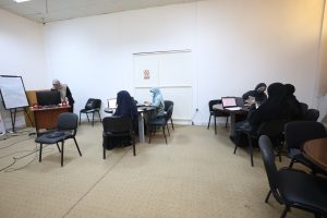 DE802954-7D6D-4AE8-A9F2-6397840E6568 - جامعة سبها | Sebha University Group of women wearing hijabs working on laptops around round tables in a classroom, with a presenter at a desk on the left.