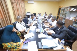 DE9A92B1-7303-4BEF-984E-A04B06409909 - جامعة سبها | Sebha University Group of professionals in a meeting around a long conference table in a bright office, with papers, bottles, and notebooks scattered about.