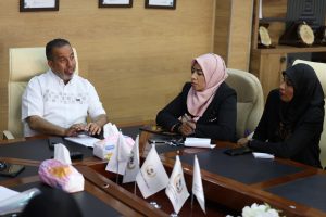 Three professionals sit around a wooden conference table: man on the left speaking, woman in a pink hijab in the center, and a woman in a black hijab on the right.