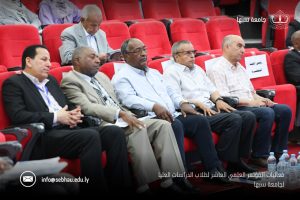 E48F76AC-3946-462F-B149-91D2EDDF5357 - جامعة سبها | Sebha University Group of men in business attire seated in red theater seats at a formal conference, Sabha University appearance visible in the corner, with contact email info@sebha.edu.ly at bottom left.