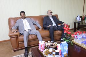 E8FCB78C-DD2A-42B6-9458-57CF60AE614D - جامعة سبها | Sebha University Two men in suits sit on a brown leather sofa in an office, with snacks and drinks on a table in front.