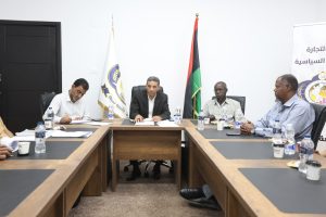 ED812E33-C29A-48D3-BE4E-C84724D79643 - جامعة سبها | Sebha University Group of men seated around a conference table in a formal meeting, with flags and an Arabic banner in the background.