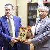 Two men in suits exchange a framed wooden plaque during an award ceremony in an office.