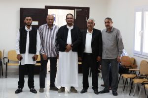 Five men stand side by side indoors, some in traditional robes and others in suits, posing for a group photo in a classroom or meeting room.