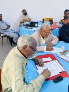 F33D0395-0E4E-4E35-B882-9E8B6BDC2D50 - جامعة سبها | Sebha University Group of men seated around a blue-clothed table signing documents, with laptops and files nearby. There are observers in the background.