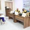 Woman in blue attire taking notes at a side table while three judges in gowns with yellow sashes sit at a panel, with a large Arabic banner behind them.