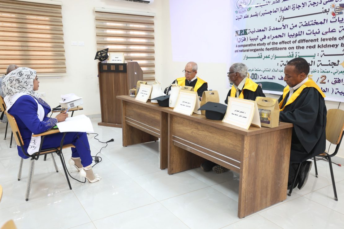 Woman in blue attire taking notes at a side table while three judges in gowns with yellow sashes sit at a panel, with a large Arabic banner behind them.