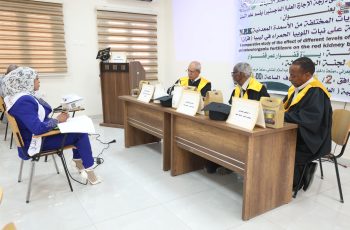 Woman in blue attire taking notes at a side table while three judges in gowns with yellow sashes sit at a panel, with a large Arabic banner behind them.