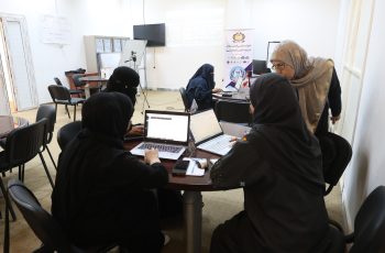 Women in hijabs seated around a table with laptops in a classroom or conference room, collaborating on a project.