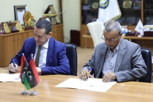 F450FD0A-3A6B-41F3-A428-3690F4B4FE06 - جامعة سبها | Sebha University Two men in suits signing documents at a conference table, with Libyan flag foregrounds.
