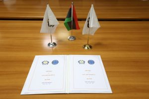 F4E83B37-1525-4169-A85A-8953D81FDD7D - جامعة سبها | Sebha University Three small flags (white, Palestinian, white) stand on a wooden desk above an open binder with official logos and text in Arabic/English.