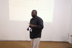 F6D564BD-1F0E-47DA-82F0-CDB2CD326DE3 - جامعة سبها | Sebha University Man in a dark shirt giving a presentation, holding papers, with a projected slide in Arabic on the wall behind him.