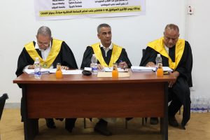 Three men in academic gowns with yellow sashes sit at a table with papers and drinks during a formal event, a banner with Arabic text behind them.