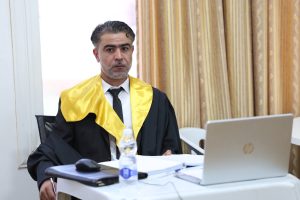FA830979-5B16-48BB-ABBA-84B8548FF827 - جامعة سبها | Sebha University Man in a black graduation gown and yellow stole sits at a desk with a laptop, notebook, and water bottle nearby in a bright room.