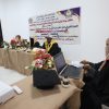 Academic panel in a ceremony hall with a banner on the wall and floral arrangements on the table, three officials in gowns and yellow sashes signing documents behind the table while a person with a laptop sits at a separate desk nearby.