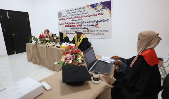 FBB2D386-7374-447E-8A6C-5CCA454EBA0F - جامعة سبها | Sebha University Academic panel in a ceremony hall with a banner on the wall and floral arrangements on the table, three officials in gowns and yellow sashes signing documents behind the table while a person with a laptop sits at a separate desk nearby.