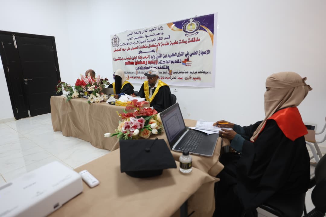 Academic panel in a ceremony hall with a banner on the wall and floral arrangements on the table, three officials in gowns and yellow sashes signing documents behind the table while a person with a laptop sits at a separate desk nearby.