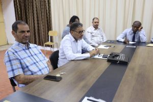 FD37AE4F-A499-450D-BA68-4BE97DBF3D61 - جامعة سبها | Sebha University Group of men seated around a conference table, talking and taking notes in a meeting room.