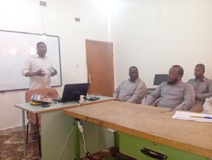 FE124F9A-FBFF-4935-B2E1-6657DF957DF2 - جامعة سبها | Sebha University Man standing at a desk giving a presentation to four men seated around a table in a classroom, with a whiteboard and laptop nearby.
