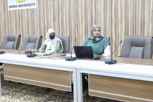 Two women in hijabs sit at a conference table with microphones in a meeting room; banner reads 'SEBHA UNIVERSITY' in the background.