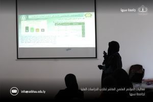 Female presenter in a hijab delivering a slide presentation in a classroom; projection screen shows charts and Arabic text, with university logos visible at the top corners.