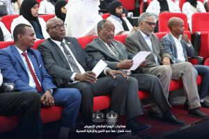 IMG_8318 - جامعة سبها | Sebha University Front-row of formally dressed men in suits sitting in red auditorium seats at a conference, with medical staff and hijab-wearing attendees in the background.
