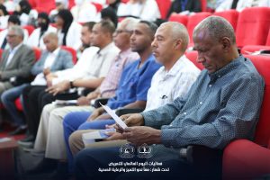 IMG_8328 - جامعة سبها | Sebha University Audience seated in a red-carpeted auditorium, foreground man reads a paper while others listen attentively during a conference event in Arabic context.