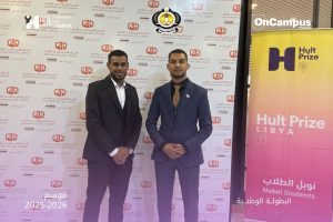 Two men in suits pose for a photo at a formal event, with a backdrop covered in logos and a Hult Prize Libya banner on the right.