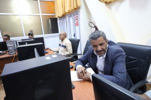Office scene with three men at computer desks; foreground man in a suit writes on a notepad while smiling at the camera.