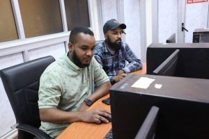 Two men seated at a wooden desk in front of computer monitors in an office, focused on their screens.