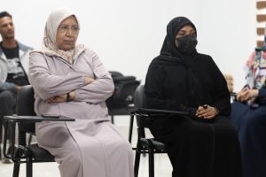 Two women in hijabs sit in a conference room; left wears a light lavender outfit with glasses, right wears a black hijab and mask.