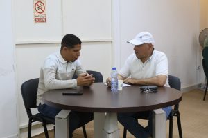 Two men sit at a round table in a plain room, talking; water bottle, cup, notebook, and keys on table nearby.
