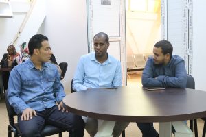 Three men sit around a dark round table in a meeting room, talking.
