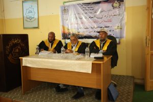 Three men in black robes with gold sashes sit at a panel desk, papers and water bottles in front, Arabic banner behind them.